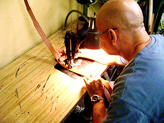 A craftsman leaning over the bench, guiding a leather belt strap under the needle of the shop's sewing machine by hand, under a work lamp.